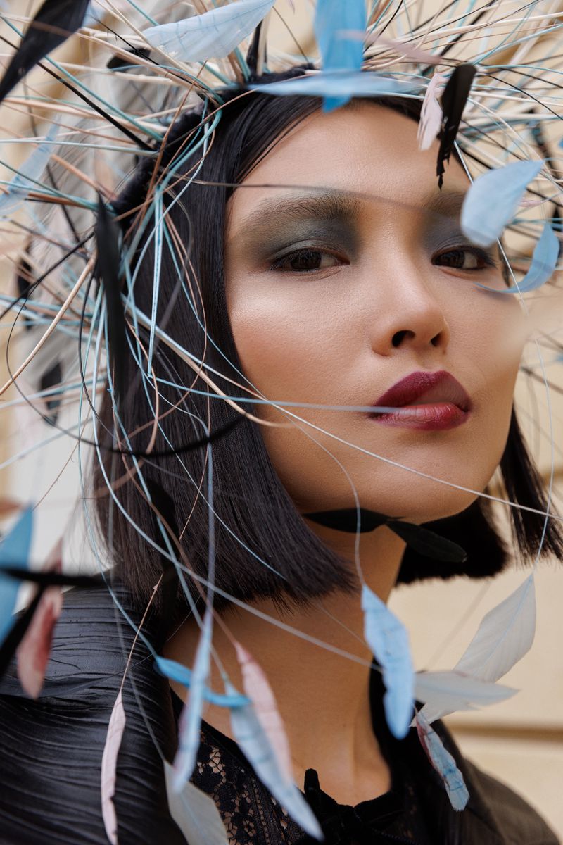 Close-up portrait of a woman with a dramatic feather headdress, smokey eyeshadow, and dark red lipstick against a neutral backdrop.
