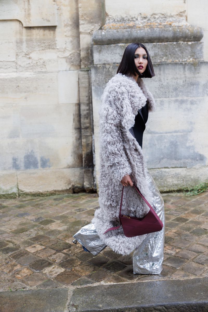 A woman walks in a grey, shaggy coat and metallic silver pants, carrying a maroon purse against a light neutral stone building.