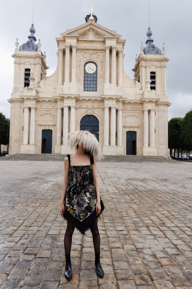 A woman in a patterned dress stands in front of a large, ornate building with a clock tower. She wears boots and opaque stockings.
