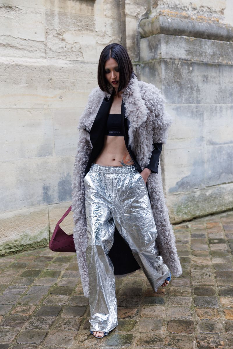 Woman wearing a grey fur coat and shiny silver pants walking on a stone path outdoors with a building visible in the background.