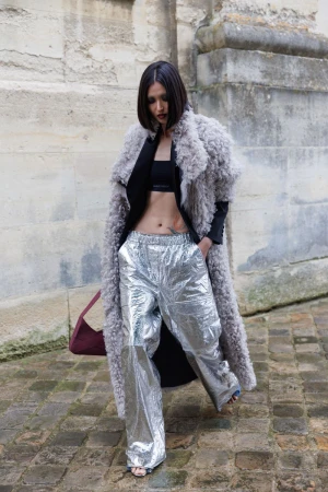 Woman wearing a grey fur coat and shiny silver pants walking on a stone path outdoors with a building visible in the background.