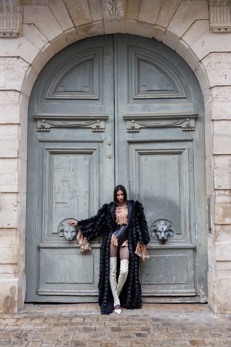 A woman in a long fur coat and thigh-high boots stands in front of a large arched stone doorway with ornate lion head details.
