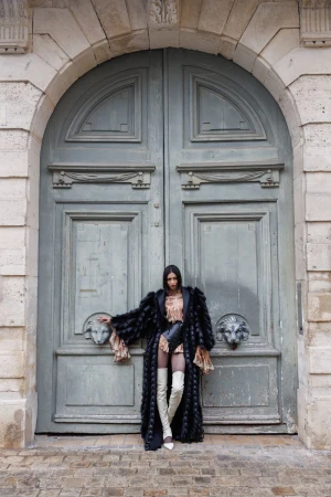 A woman in a long fur coat and thigh-high boots stands in front of a large arched stone doorway with ornate lion head details.
