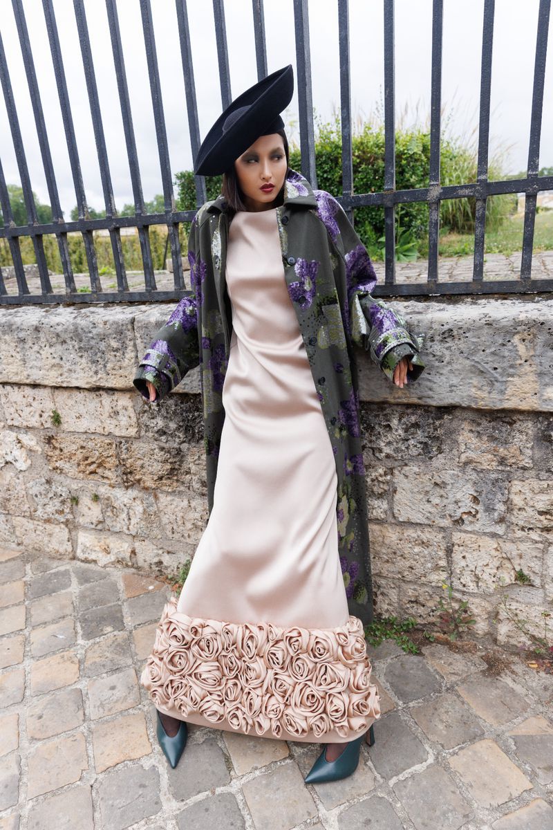 Adult female in a beige dress with flower detail, wearing a black hat and floral coat, stands against a stone wall with a metal fence.