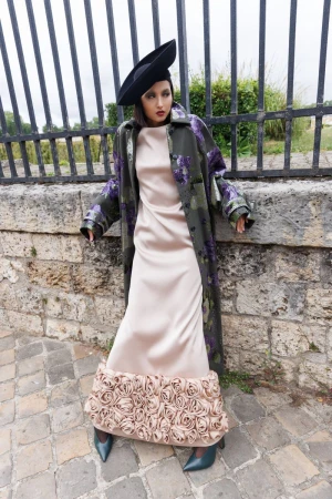 Adult female in a beige dress with flower detail, wearing a black hat and floral coat, stands against a stone wall with a metal fence.