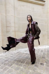 Model Cynthia Wu wearing a burgundy leather vest and wide-leg trousers while posing dynamically on cobblestone pavement.