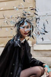 Model wearing an elaborate feather headpiece and black leather coat outdoors against a textured building wall for L'Officiel Monaco.