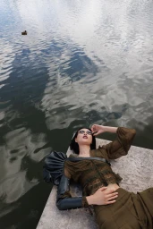 Model wearing an olive green top and trousers lying on a stone edge beside rippled water for L'Officiel Monaco.
