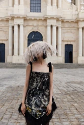 Model Cynthia Wu wearing an exaggerated feather headpiece and patterned feather-trim dress in front of a historic stone church.
