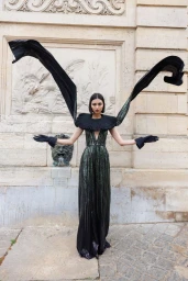 Model in a black sequin jumpsuit with dramatic winged fabric extensions and a ruffled collar standing outdoors.