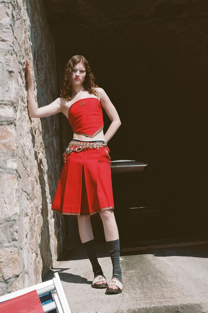 A stylish young woman poses in a red tube top and skirt with unique accessories on a stone patio in front of a dark opening.
