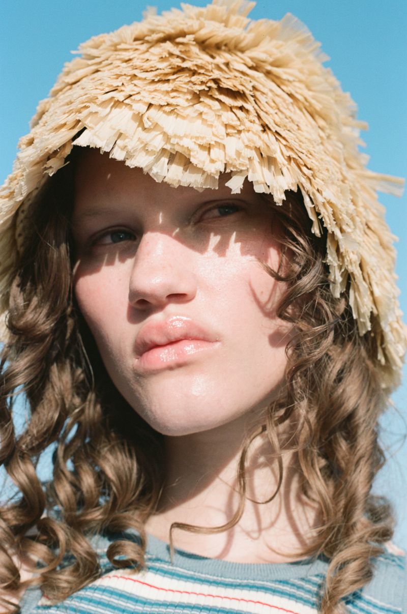 Close-up portrait of a fair-skinned adult female wearing a textured beige hat against a clear blue sky in an outdoor environment.