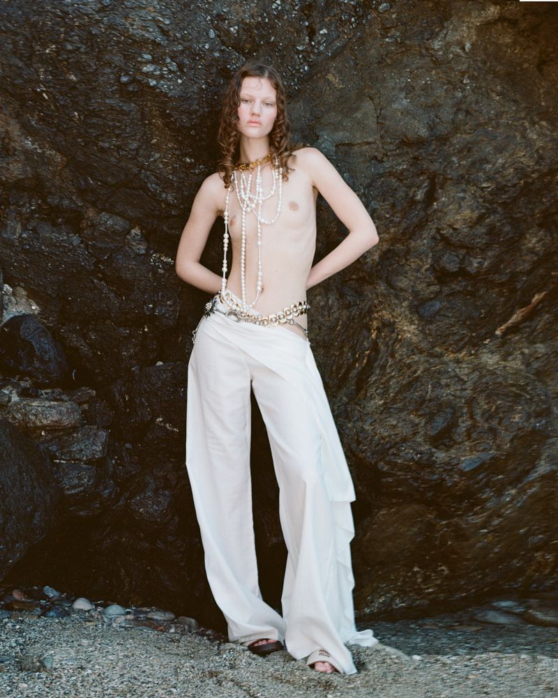 A model in a pearl necklace and white pants poses against a rock, creating a natural and fashionable outdoor shot.