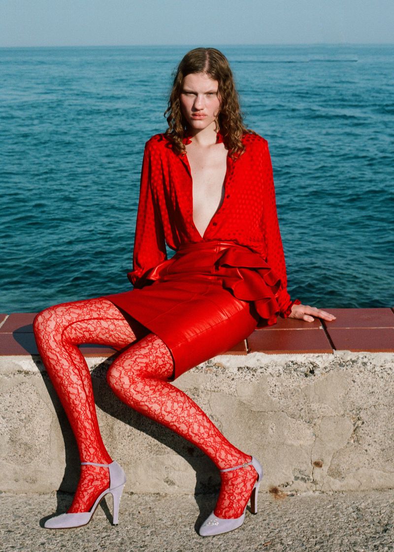 Fashion photo of a young woman in a red outfit with lace tights and lavender heels sitting against a sea background in bright daylight.