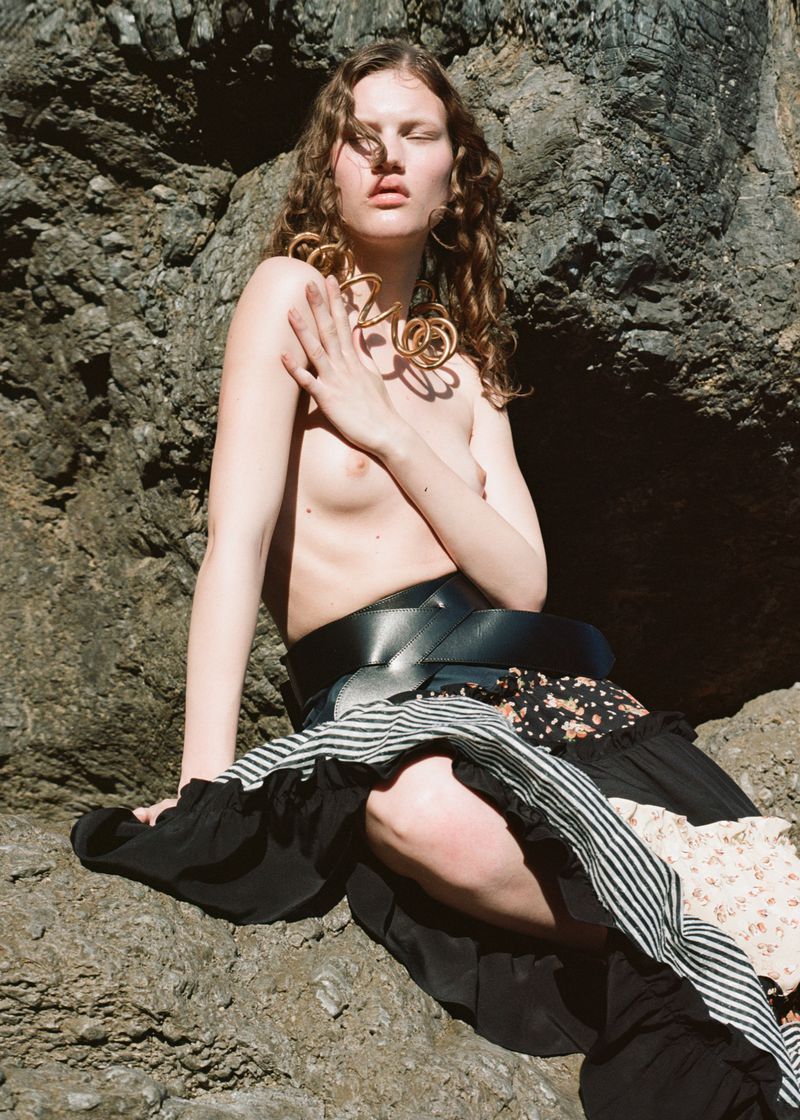 A nude woman with curly hair and gold jewelry sits on a rock in a fashion shot featuring natural light and a dark textured background.