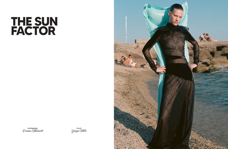 A model in a black lace dress poses on a sandy beach with an inflatable pool float, with the sea and other people in the background.