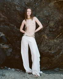 Model standing against a dark rocky coastal backdrop wearing layered pearl necklaces and loose white trousers.