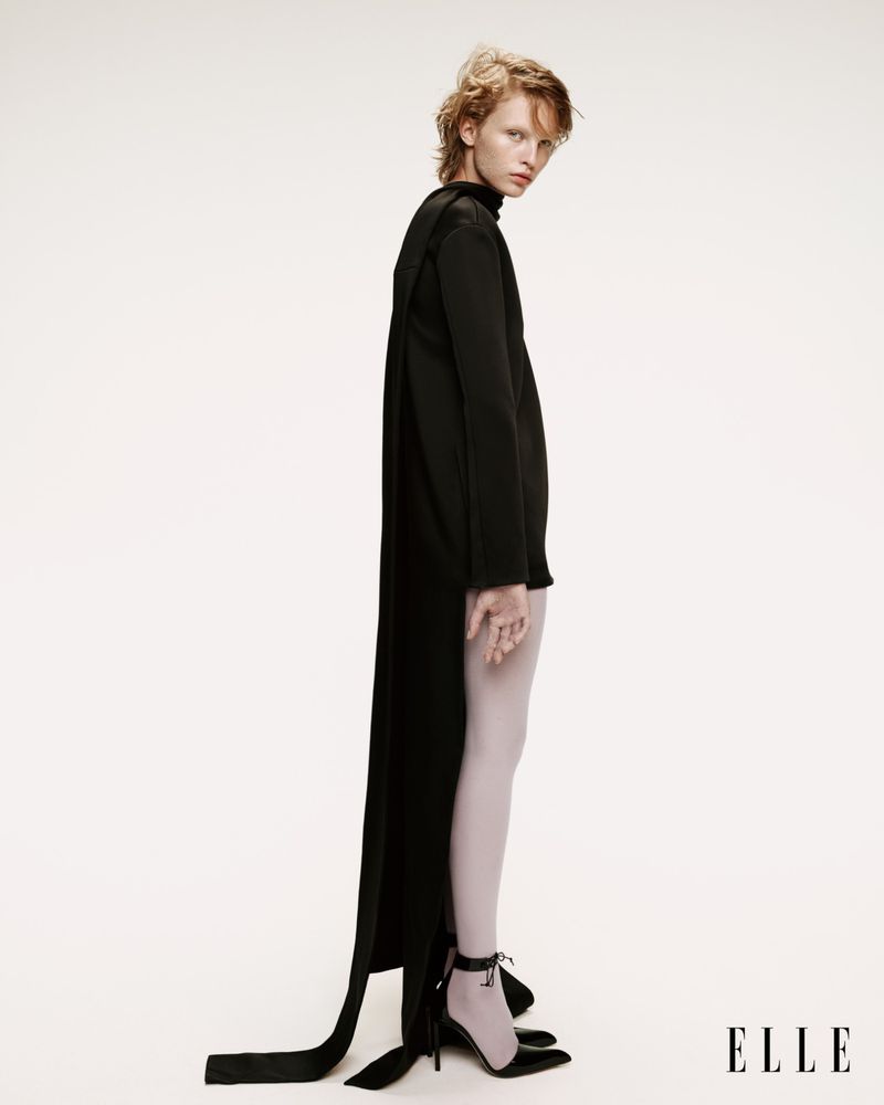 Fashion editorial shot of a woman in a black dress with white tights and black heels against a white backdrop with ELLE logo.