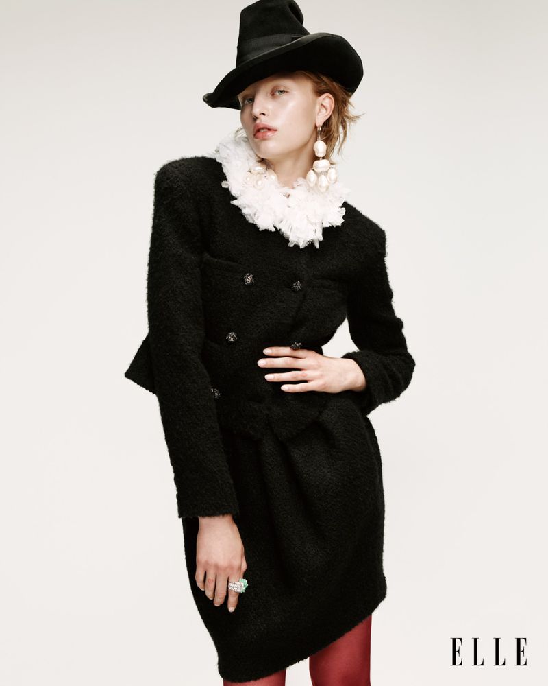 A woman poses in a black, textured jacket and skirt with a matching black hat, accessorized with white jewelry, against a neutral backdrop.