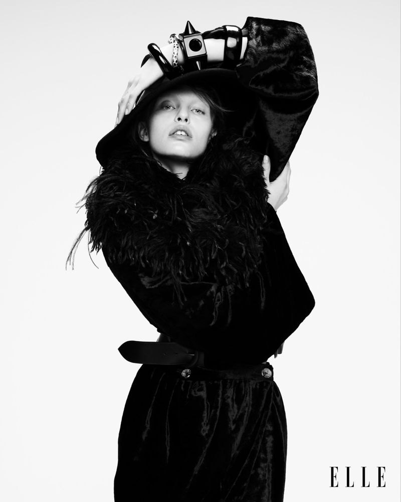 Portrait of a woman wearing a black coat with feathered trim and a hat, accessorized with bracelets, against a white backdrop.