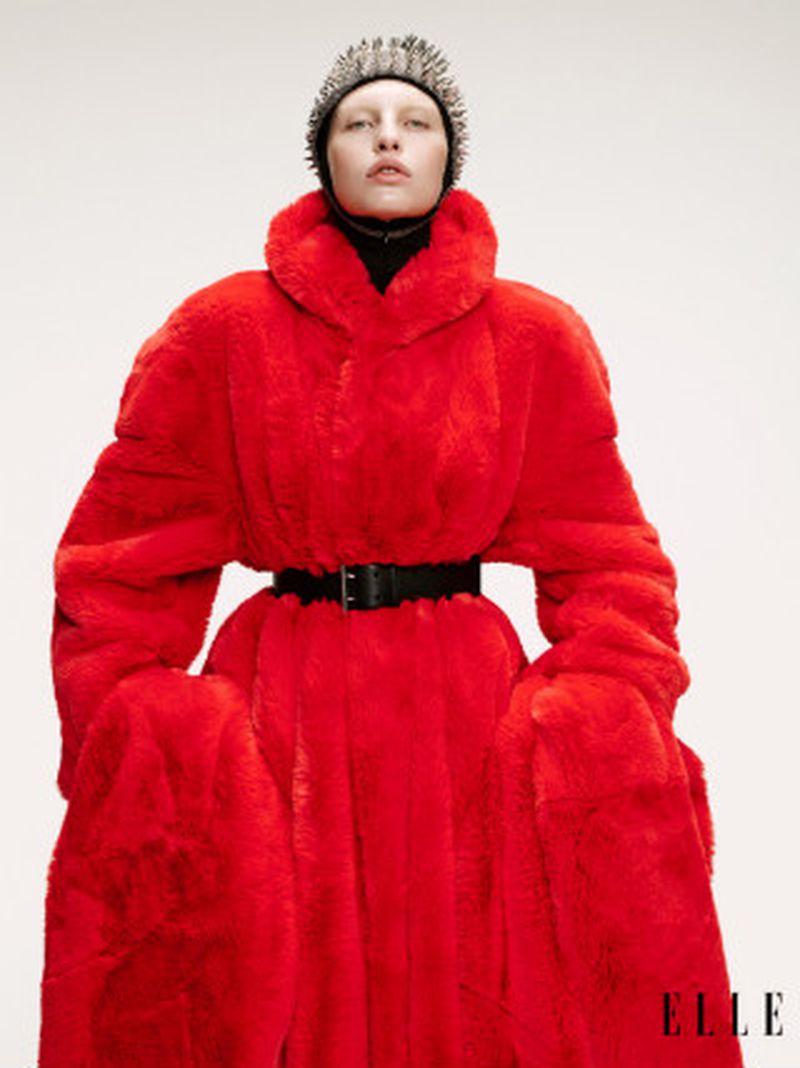 Fashion shot of a model wearing a red faux-fur coat, black belt, and spiked hat against a white background. Part of an ELLE magazine editorial.