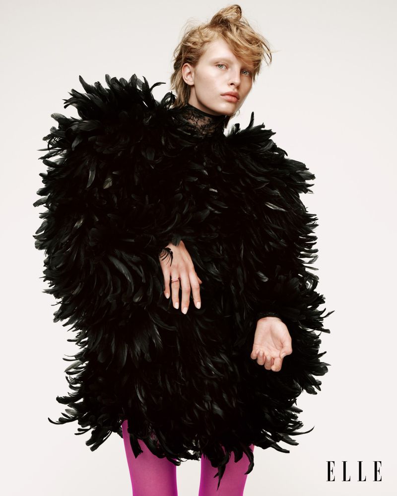 Fashion shot of a blonde woman wearing a black feather coat and pink tights against a white backdrop, Elle logo visible.