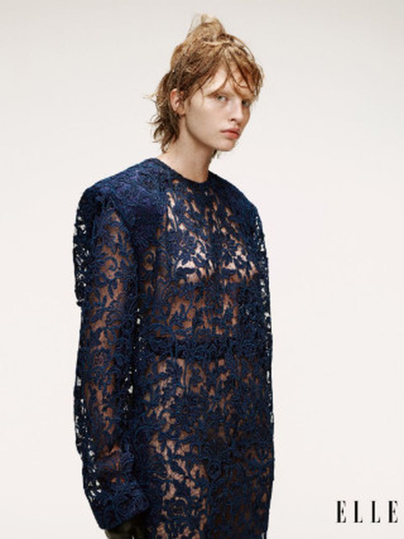 Fashion editorial shot of a fair-skinned woman with short, wavy hair, wearing a navy blue lace dress, posing in front of a white backdrop.