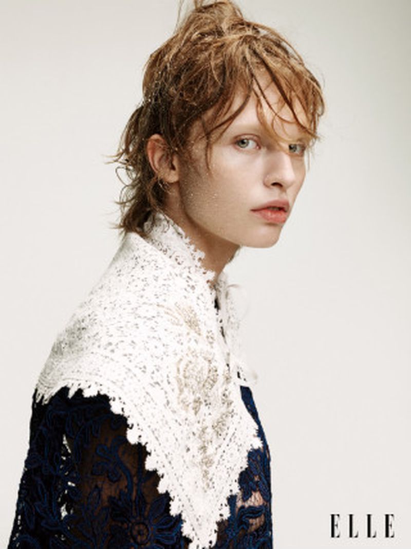 Portrait of a person with wet, styled hair, wearing a dark blue lace top with a white lace collar, against a soft white background.