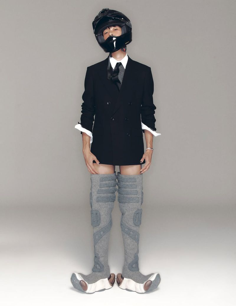 Person in black suit, tie, helmet, and gray thigh-high boots poses in a studio against a gray backdrop. Fashion and portrait photography.