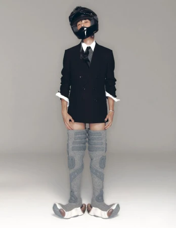 Person in black suit, tie, helmet, and gray thigh-high boots poses in a studio against a gray backdrop. Fashion and portrait photography.