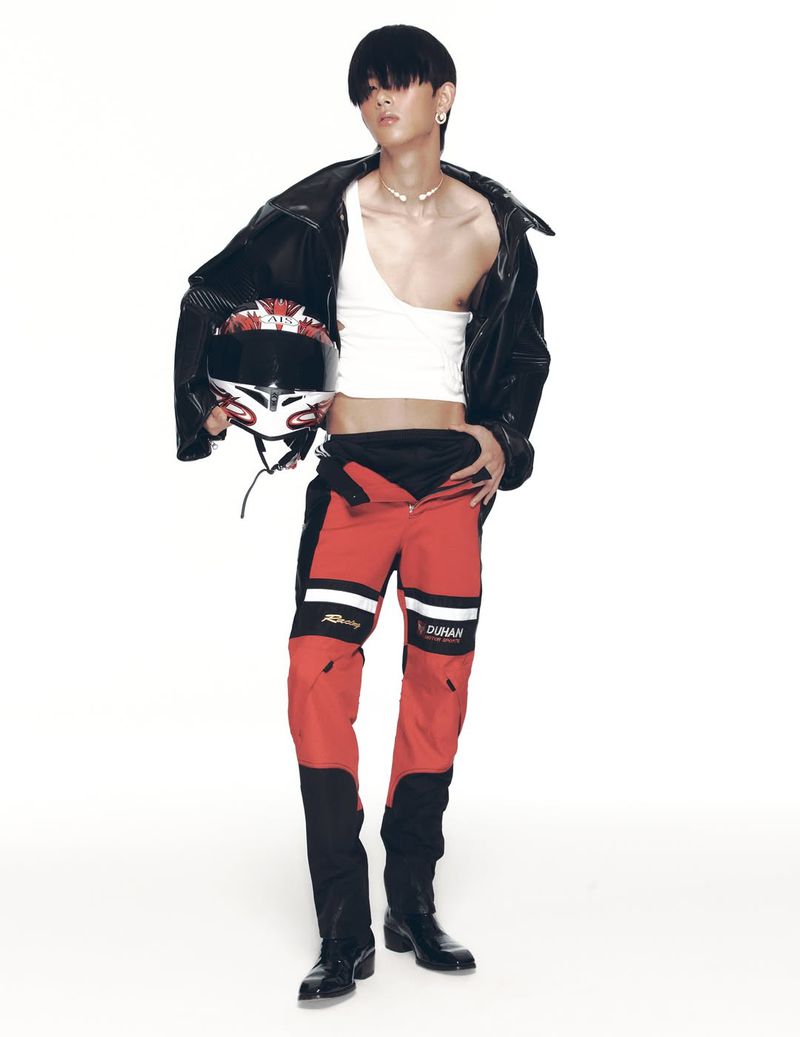 Full shot of adult male in red and black moto pants, leather jacket and white crop top, holding a motorcycle helmet on a white backdrop.