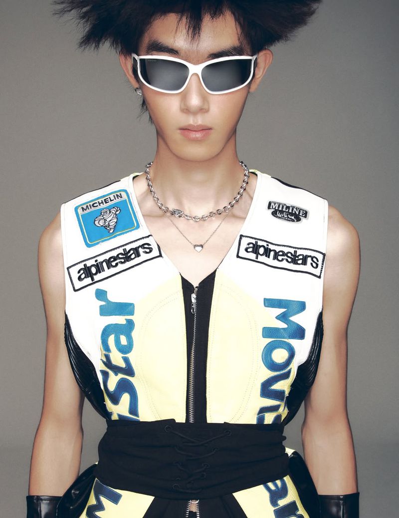 Close-up of a young adult with spiky hair and sunglasses wearing a racing-themed vest with logos and graphic designs against a gray background.