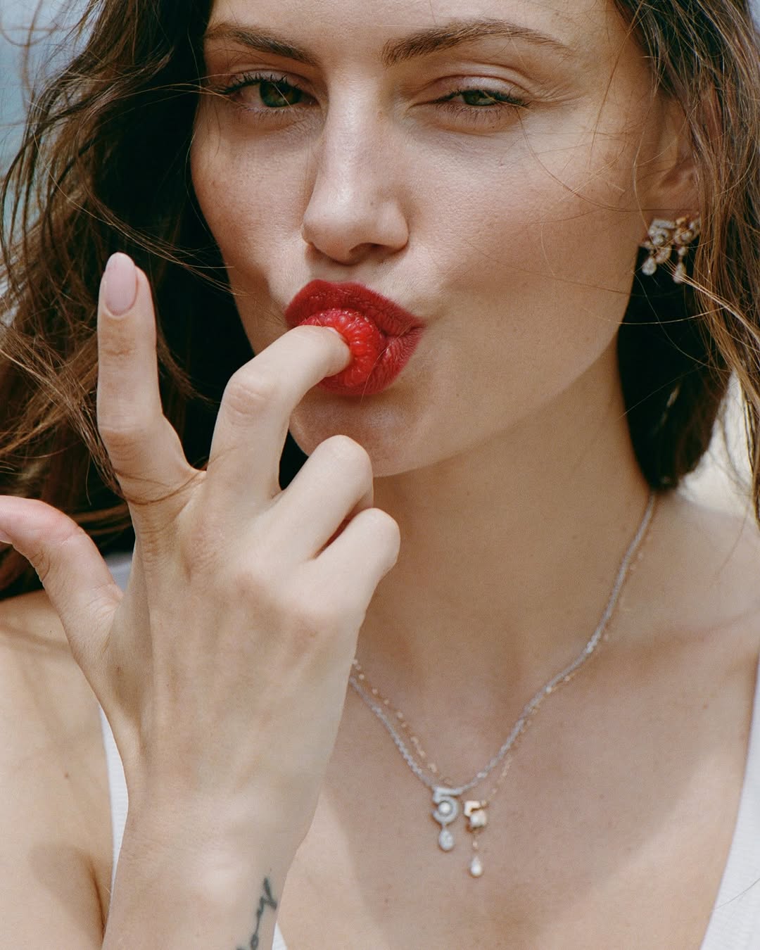 Close-up of a woman with dark hair wearing earrings and necklaces, holding a red raspberry to her red lips, with a blurred background.