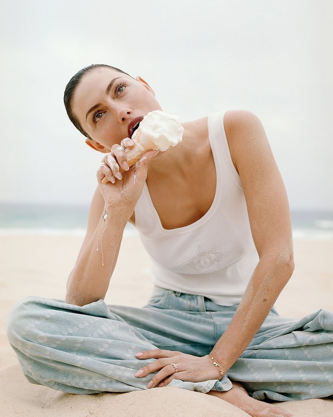 Phoebe Tonkin seated on a sandy beach wearing a white tank top and light blue denim pants while eating an ice cream cone