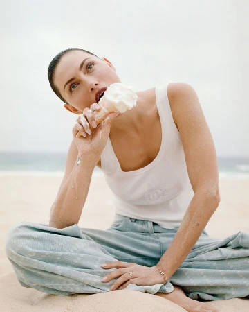 Phoebe Tonkin seated on a sandy beach wearing a white tank top and light blue denim pants while eating an ice cream cone