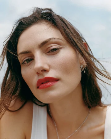 Close-up portrait of Phoebe Tonkin wearing red lipstick and a white top under an overcast sky.