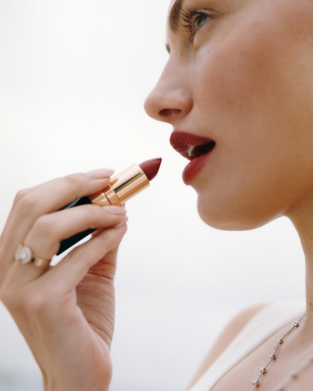 Close-up of a woman applying dark red lipstick, showcasing her hand with a ring and a delicate necklace against a bright background.