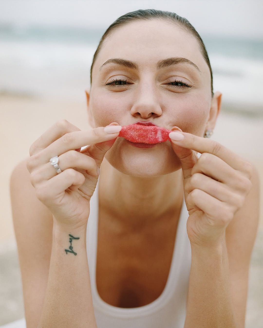 Woman poses with a candy as fake lips on a beach with a white tank top and a visible tattoo, creating a playful and fun image.