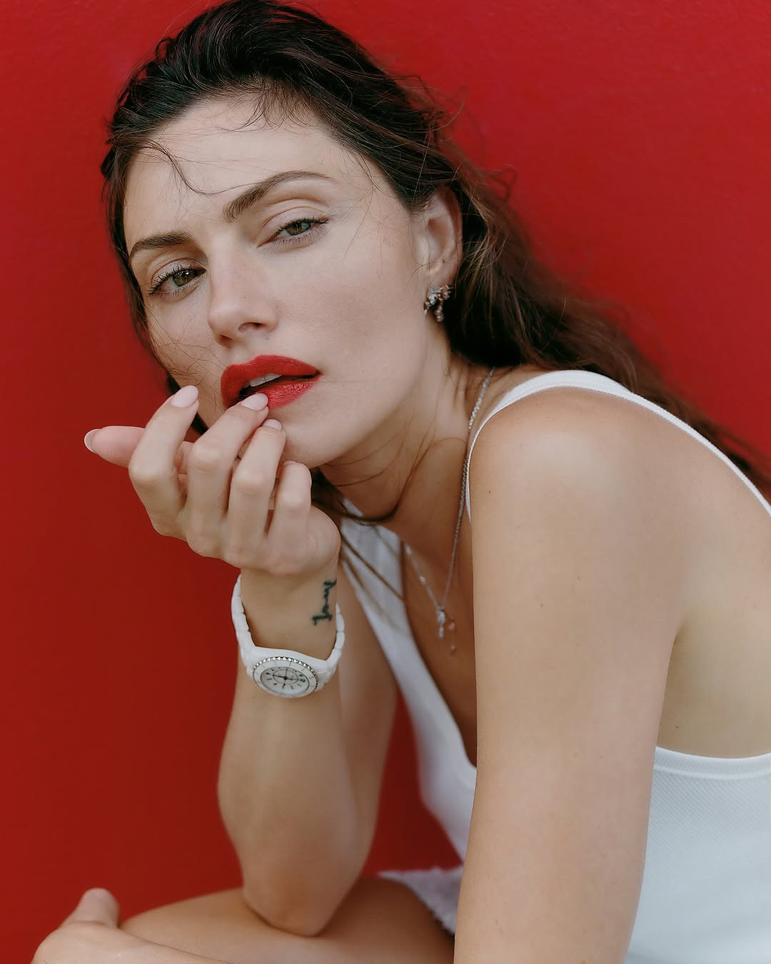 Close-up portrait of an adult woman with red lipstick, wearing a white top and wristwatch, against a solid red background in a studio.