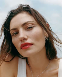 Close-up portrait of Phoebe Tonkin wearing red lipstick and a white top under an overcast sky.