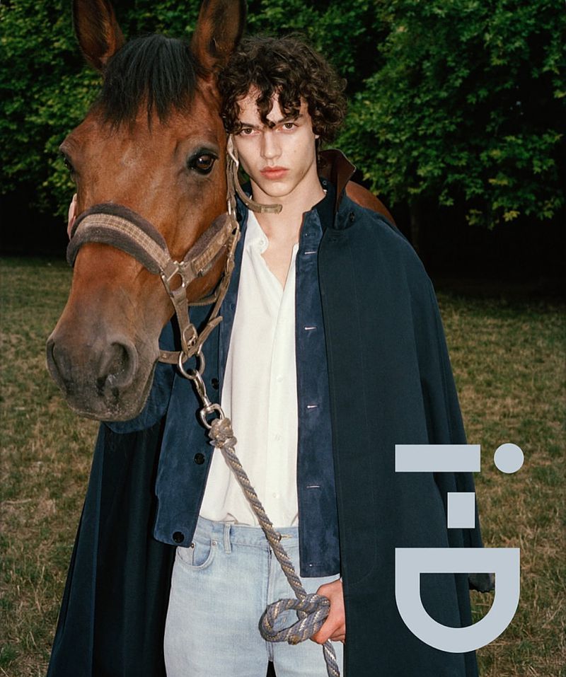 Young man poses with a brown horse outdoors, wearing a blue jacket, white shirt, and jeans in a fashion editorial shot.