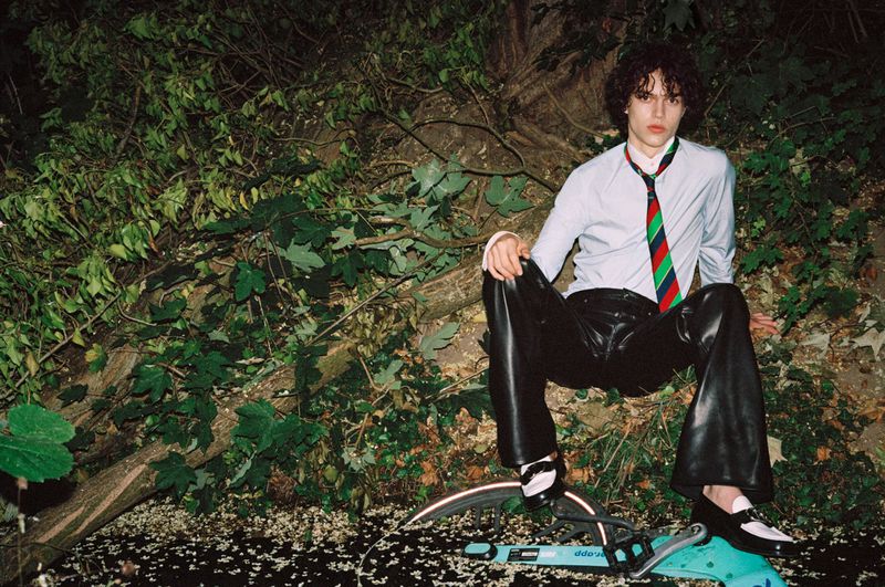 Adult male with curly hair sits on a bicycle wearing a light blue dress shirt, colorful tie, and black leather pants in an outdoor setting.