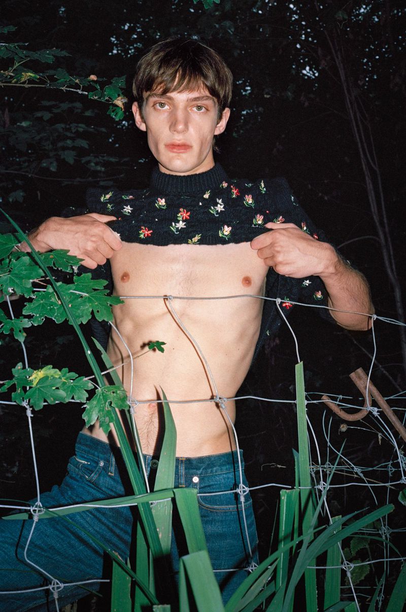A slender adult male poses outdoors, partially exposing his torso in front of foliage, a wire fence, and a dark, wooded background.