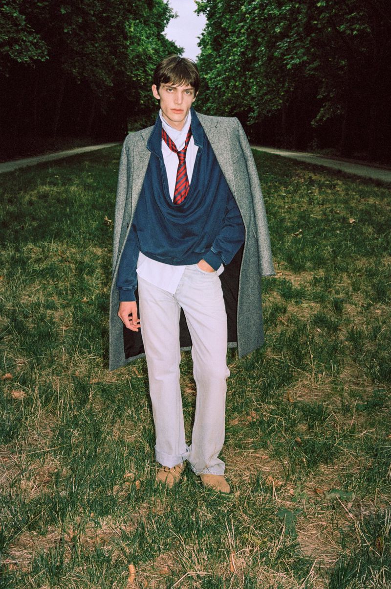 Model Cruz Kahlbetzer in a grey overcoat, navy cowl-neck top, and red striped tie standing on a grassy park path.