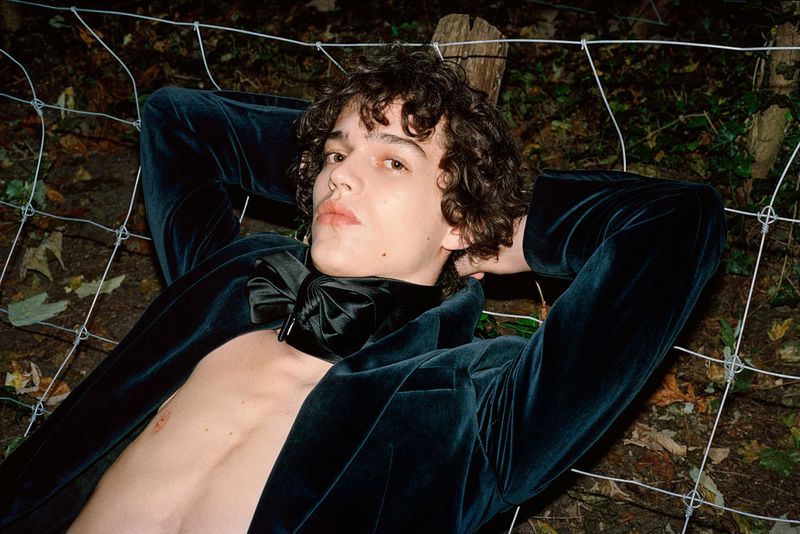 Young adult with curly hair lies in repose wearing a dark teal velvet suit jacket and oversized bow tie against a blurred foliage backdrop.