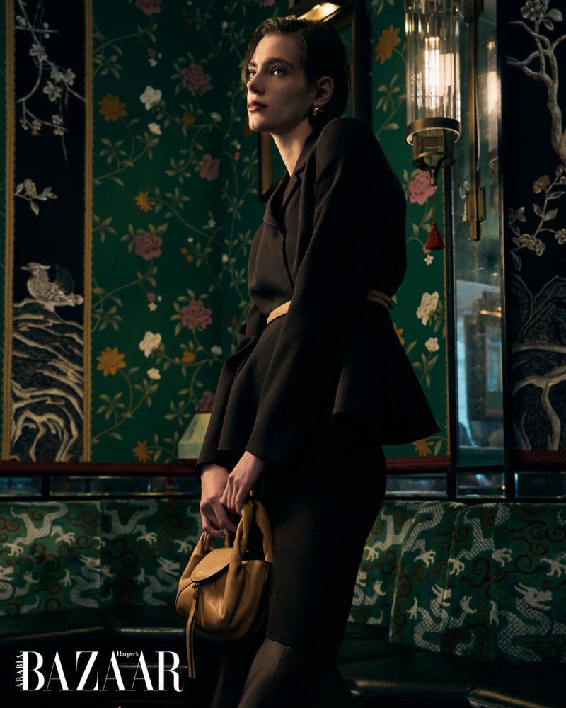 A woman in a dark business suit stands indoors holding a tan purse, with floral wallpaper and decorative lighting in the background.