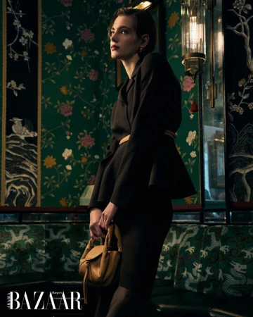 A woman in a dark business suit stands indoors holding a tan purse, with floral wallpaper and decorative lighting in the background.