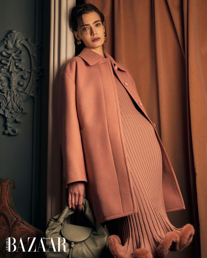 Fashion editorial shot of a model in a pink coat and dress holding a purse against a textured wall and draped fabric.