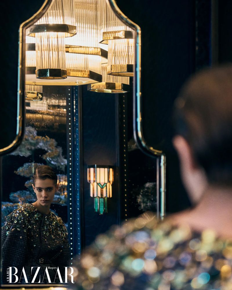 A woman in a sequined top looks into a black-framed mirror with a decorative chandelier, featuring a magazine logo at the bottom.