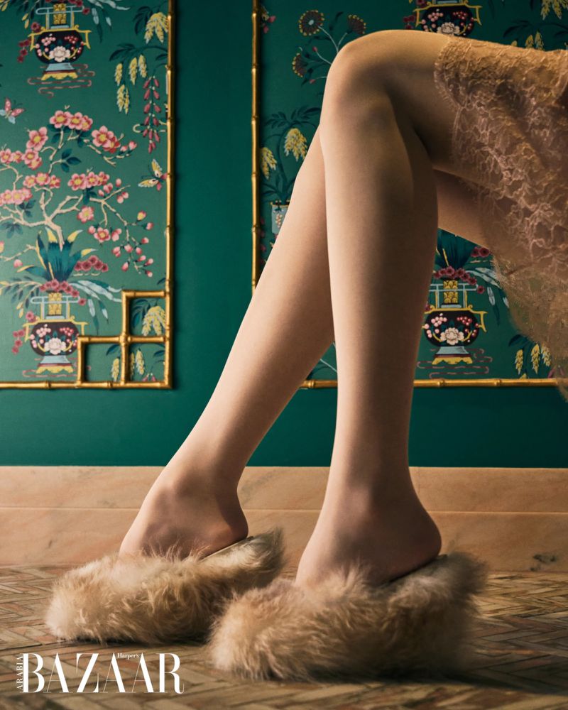 Woman's legs and feet wearing fur slippers, set against a green floral wallpaper, showcasing fashion footwear indoors.